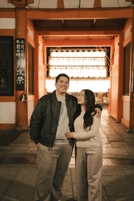 Smiling couple standing in front of traditional Japanese wooden architecture with lanterns at night