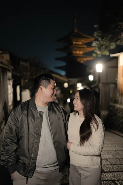 Couple smiling at each other on a street at night with a pagoda