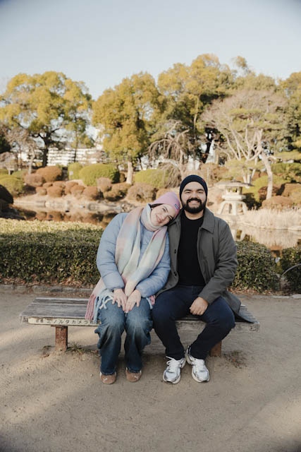 Couple sitting on a wooden bench in a garden with trees and a pond behind them