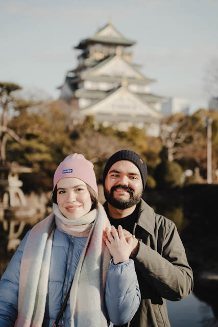 Smiling couple in winter beanies and coats clasping hands in front of a traditional Japanese castle