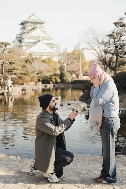 Man proposing to surprised woman in pink beanie near pond with castle background