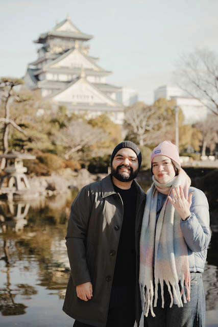 Couple in winter coats posing in front of Osaka Castle with engagement ring visible