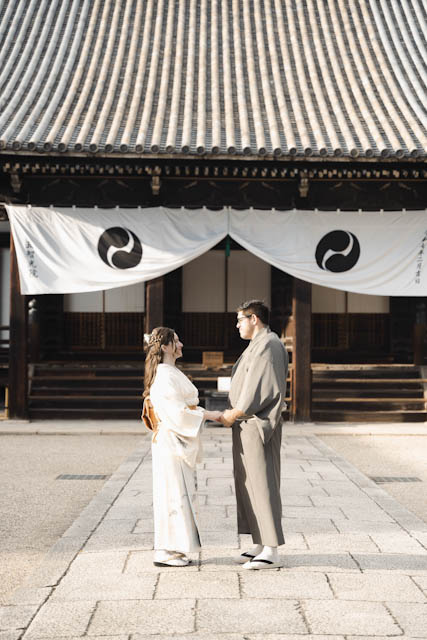 Couple in traditional Japanese clothing holding hands in front of a temple building with white banners