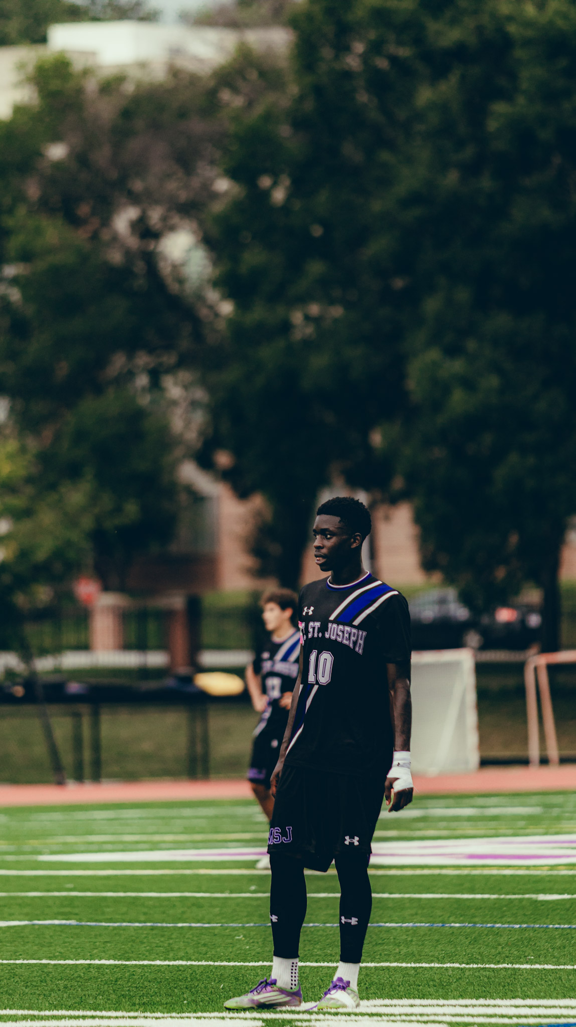 A young male soccer player wearing a dark jersey with the number 10 stands on the field, looking towards the game. Another player is in the background, with trees and a school building visible.