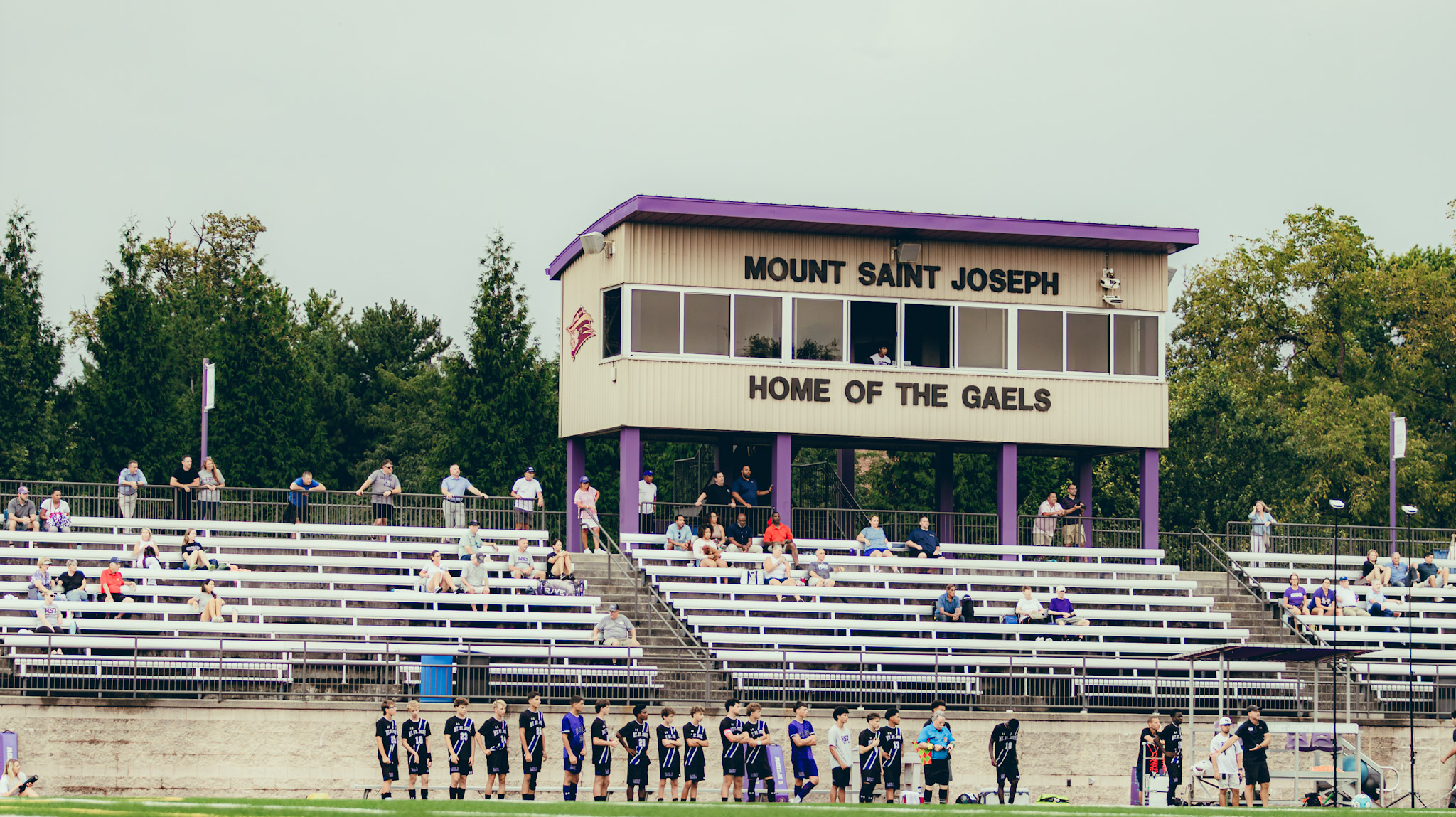 A soccer field at Mount Saint Joseph, featuring a scoreboard marked 'HOME OF THE GAELS' and spectators across the bleachers, with players lined up on the side.
