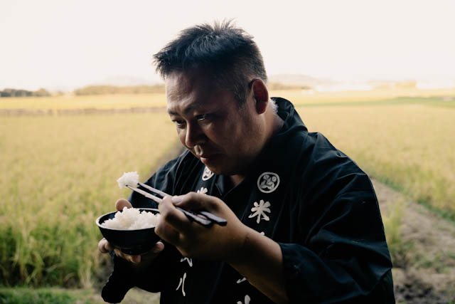 田んぼの背景で黒い作業着を着て白米を食べる男性のポートレート