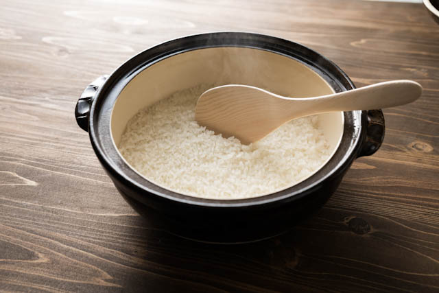A steaming black ceramic rice bowl with a wooden rice paddle on a wooden table.