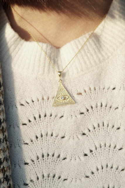 Gold triangular pendant with eye design on a white knitted sweater