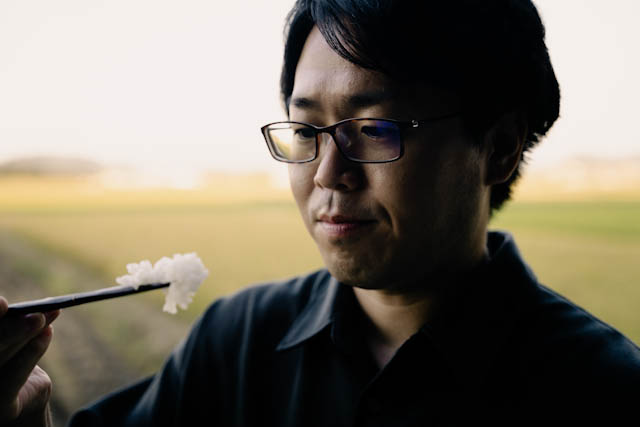 自然の風景を背景に、男性が箸で白い食べ物を食べる様子を捉えたポートレート写真