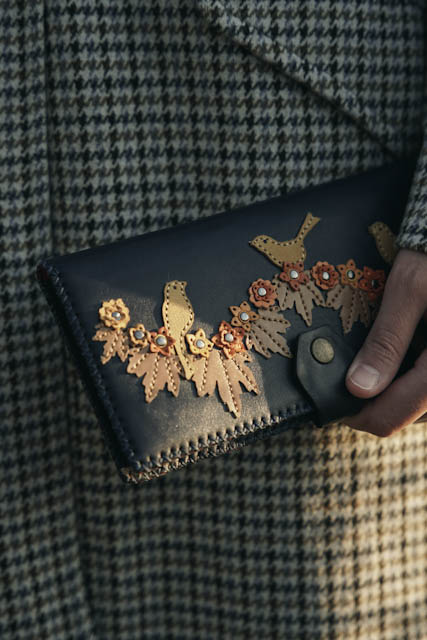 Hand holding a dark leather clutch with tan bird and flower cutouts against a houndstooth coat