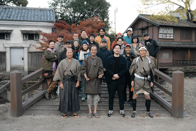 日本家屋と紅葉を背景に、和服姿のキャストとスタッフが木橋で集合写真を撮影している