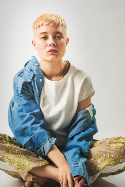 Person with short blonde hair wearing denim shirt and embroidered skirt sitting cross-legged