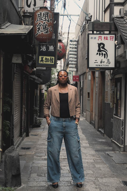 Woman in beige blazer and jeans standing in a narrow Japanese alley with traditional lanterns