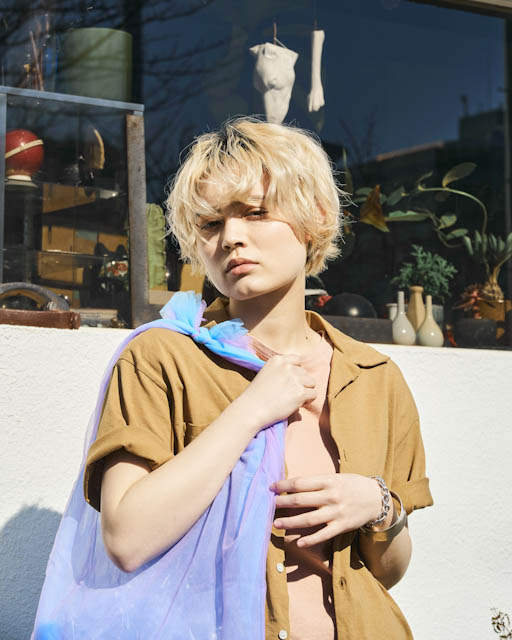 Short-haired person wearing brown shirt and blue scarf outdoors