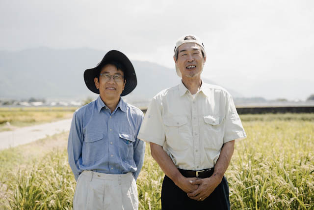 Two smiling men standing in a rice field with mountains in the background