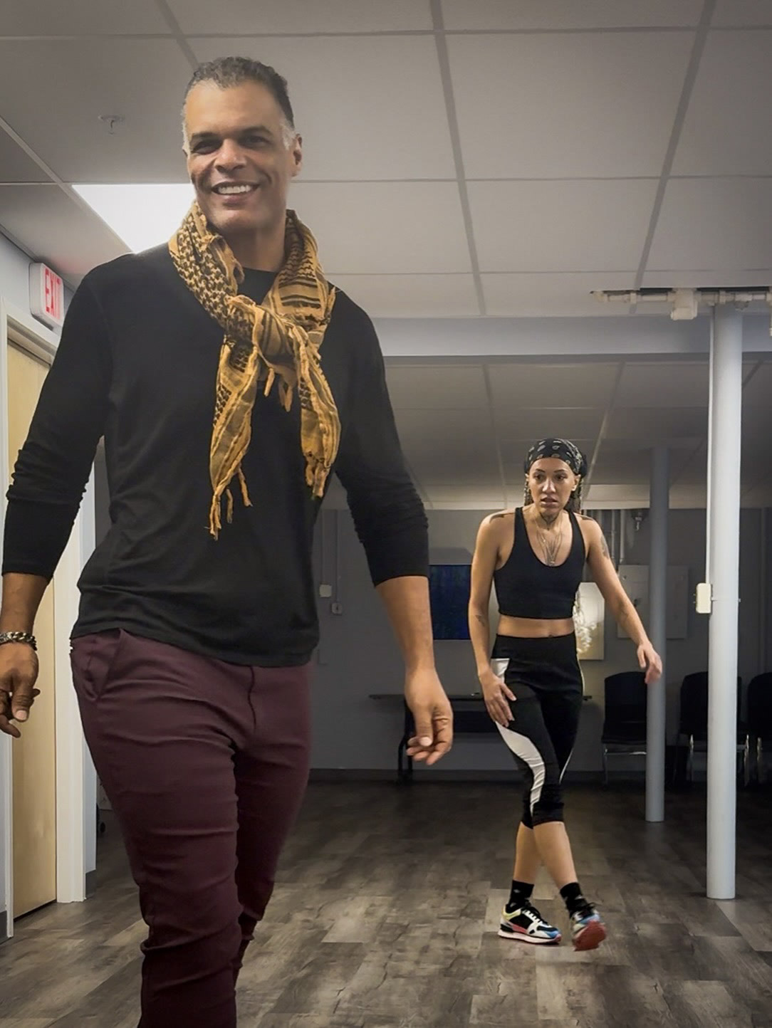 Kristopher wearing a black shirt, scarf, and dark red trousers in a studio.
