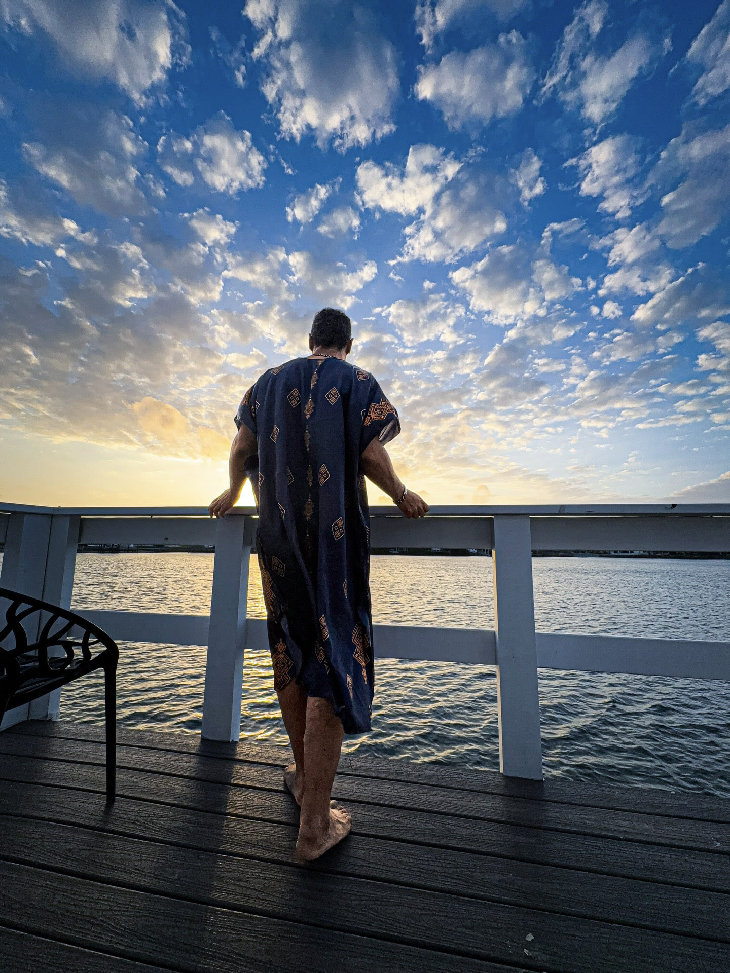 Kristopher standing at a railing beside the water in a flowing patterned garment.
