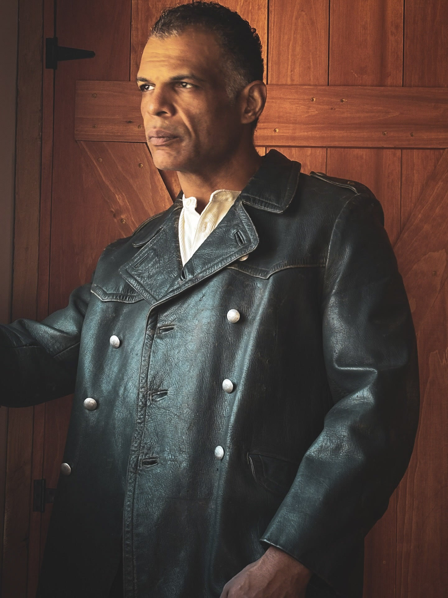 Kristopher wearing a vintage dark leather coat beside a wooden door.