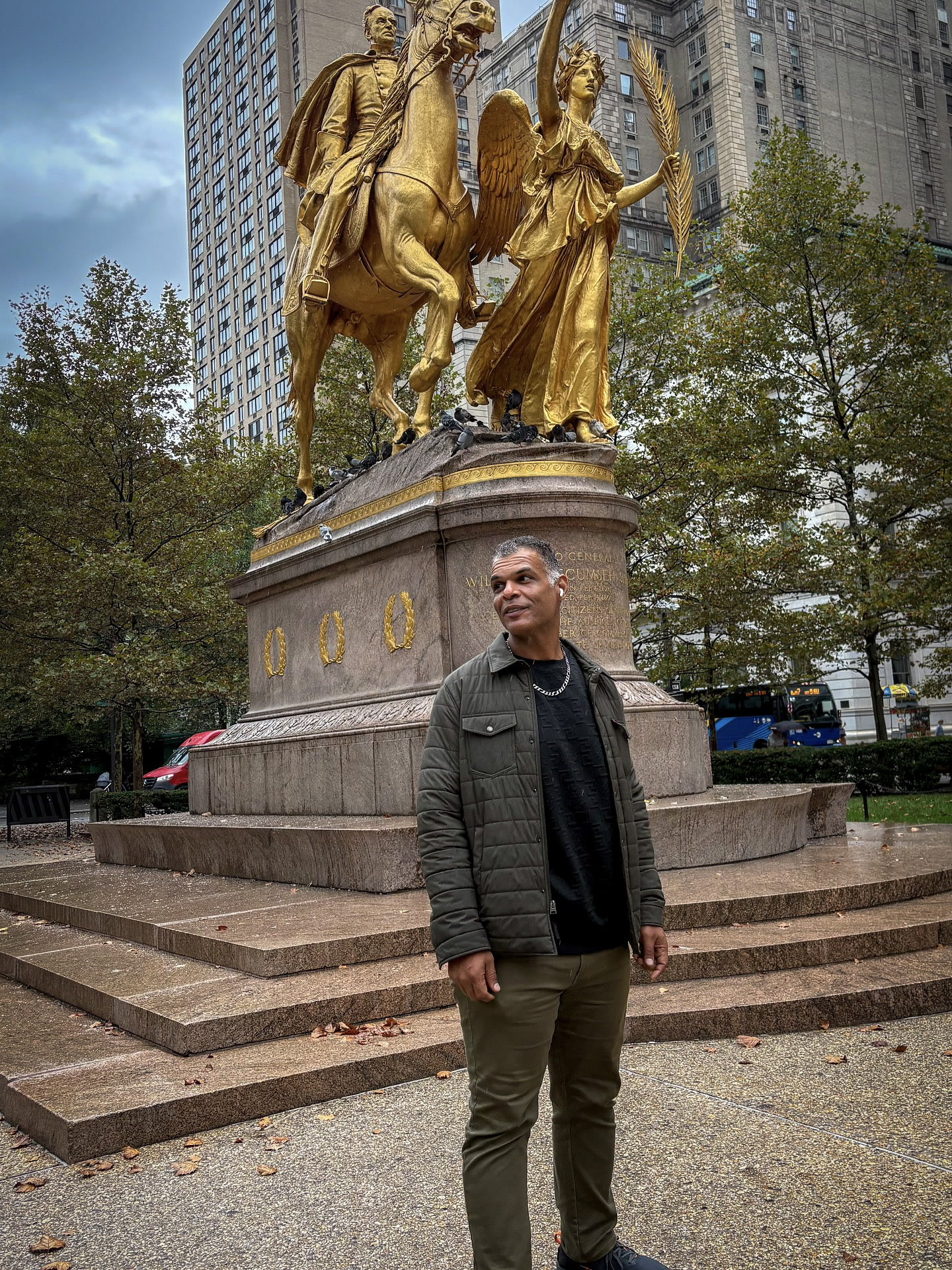 Kristopher standing in an olive jacket beside a monument in New York.