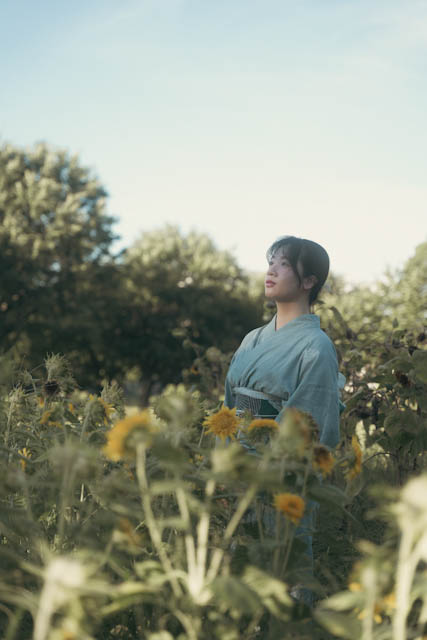 Woman in light blue kimono standing among sunflowers looking up