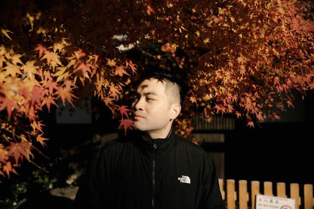 Man in black jacket standing under red maple leaves in sunlight