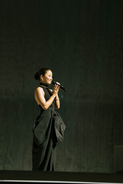 Woman in black dress holding microphone on stage under spotlight