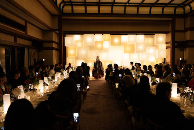 Performer in traditional costume walking down aisle between seated guests and glowing lanterns