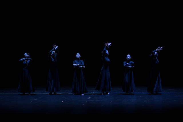 Dancers in long black dresses performing on a dark stage with blue lighting
