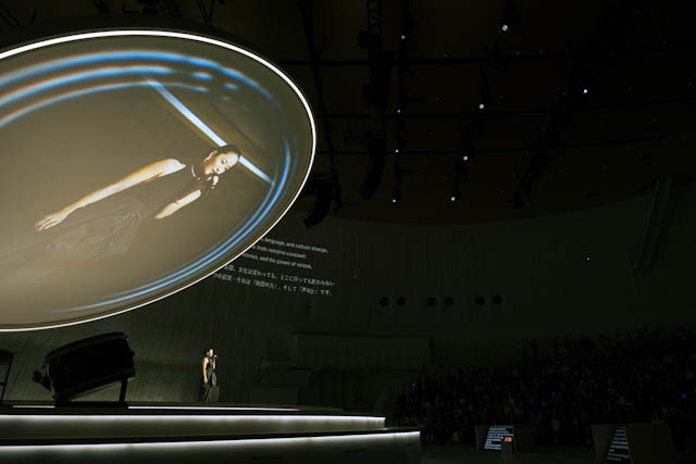 Speaker standing on stage beside piano with large circular projection overhead