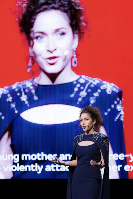 Woman in navy blue dress speaking on stage with red screen background